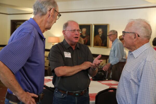 Three older men talk near a table in a room with framed portraits at First Presbyterian Baton Rouge.