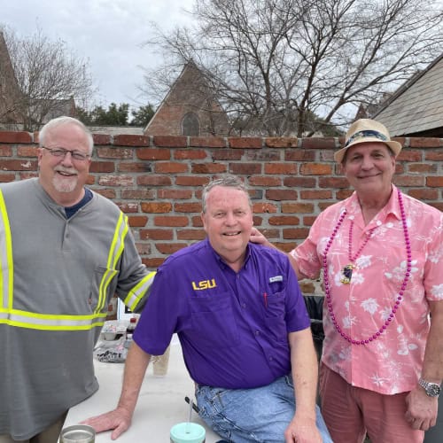 Three men smile outdoors by a brick wall, one in a safety shirt, at First Presbyterian Baton Rouge.