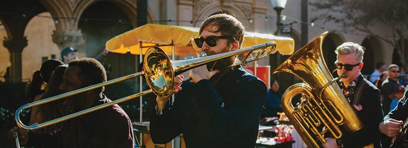 A group of musicians plays brass instruments outdoors at First Presbyterian Baton Rouge; one plays trombone, another tuba—both wear sunglasses.
