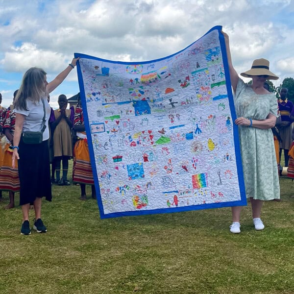 Two women hold up a colorful handmade quilt outdoors as members of First Presbyterian Baton Rouge stand behind them in traditional clothing.