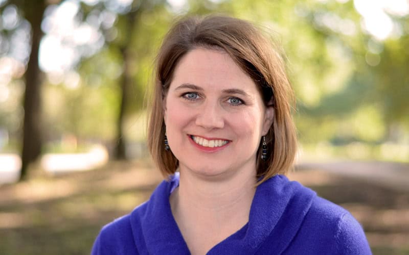 Woman with short brown hair in a blue top, smiling outdoors near first Presbyterian Baton Rouge with trees and sunlight in the background.
