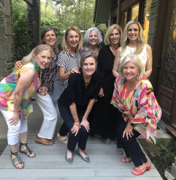 Eight women pose and smile together outdoors on a wooden deck near a house, surrounded by greenery and trees in the background.