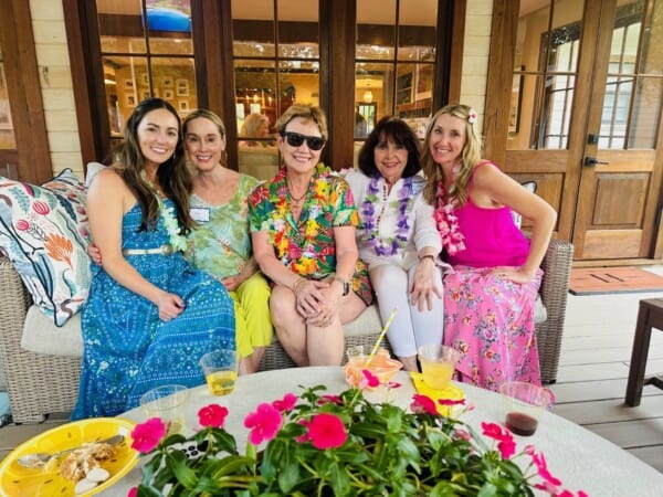 Five women sit closely together on an outdoor couch, smiling, with drinks and flowers on a table in front of them.
