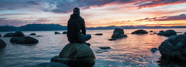 A person sits on a large rock by a lake, watching a sunset over mountains, reminiscent of the peace at First Presbyterian Baton Rouge.