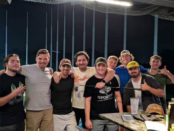 Nine men stand closely together at a driving range at night, smiling at the camera with golf clubs and drinks visible in the foreground.