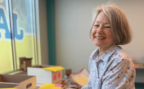 A woman with gray hair smiles while sitting at a table with boxes marked "You Did It Again" and colorful papers near a window.