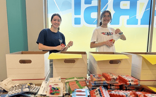 Two people stand behind boxes, sorting school supplies with care—You Did It Again in this brightly lit room.