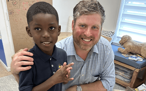 An adult mentor and a child pose together indoors, both smiling warmly at the camera—reflecting the spirit of Kids Hope USA.