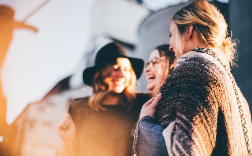 Three women from the Women's Ministry stand close together outdoors, smiling and laughing with sunlight shining in the background.