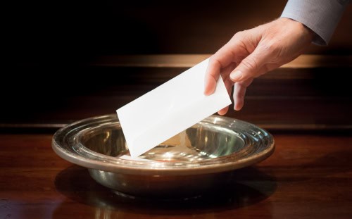 A hand places a white envelope, possibly containing Quarterly Statements, into a silver collection plate on a wooden surface.