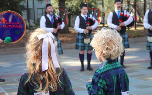Two children with name tags watch four adults in kilts play bagpipes at a Scottish event outdoors during the day.