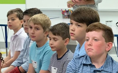 Six boys sit in a row indoors, wearing striped or solid shirts and name tags, looking attentive at their Elementary Sunday School class.
