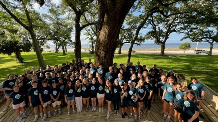 A large group in matching shirts poses outdoors at the EPiC Middle School Retreat, beneath a big tree with grass and a road in the background.