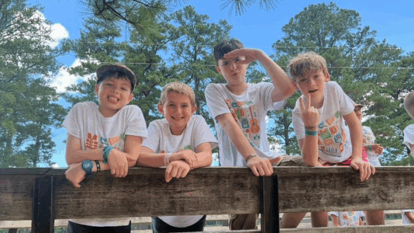 Four boys in white t-shirts lean on a wooden railing at Lake Forest Ranch, surrounded by tall trees and a blue sky.