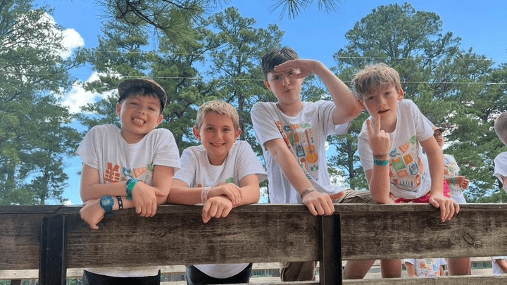 Four boys in white t-shirts lean on a wooden railing at Lake Forest Ranch, surrounded by tall trees and a blue sky.