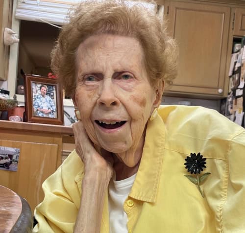 Member Spotlight: Marshall Ann Bourgeois, an elderly woman in yellow, sits at a table with a framed photo and kitchen cabinets behind her.