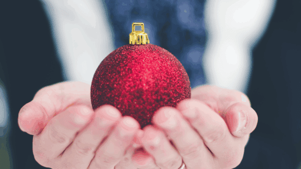 A person holding a red glittery Christmas ornament, ready to fill Christmas Bags for Donations; background softly blurred.