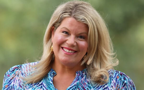 Lindsay Stratman, with long blonde hair, smiles at the camera in a blue patterned blouse, set against a blurred green background.