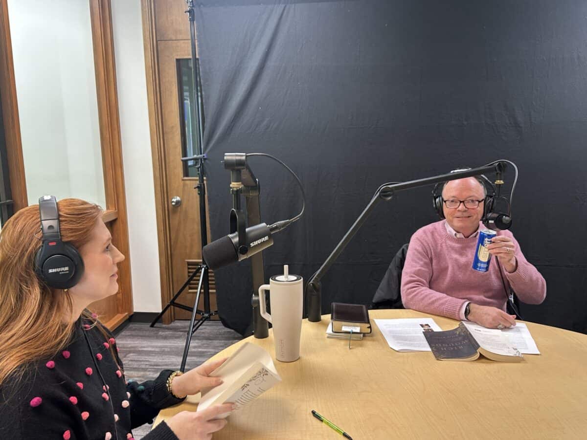 Two people wearing headphones are recording a podcast at a table with microphones, drinks, papers, and books in front of them.