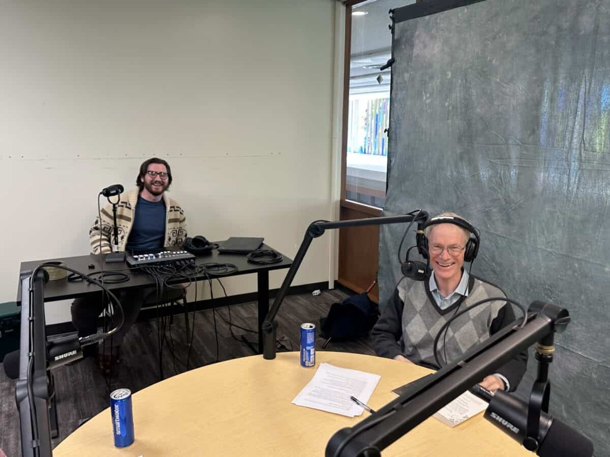 Two men wearing headphones sit at desks with microphones, audio equipment, papers, and energy drinks in a podcast recording setup.