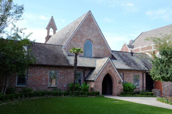 A red brick building with a steep roof, arched doorway, and small trees in front, set against a blue sky with light clouds.