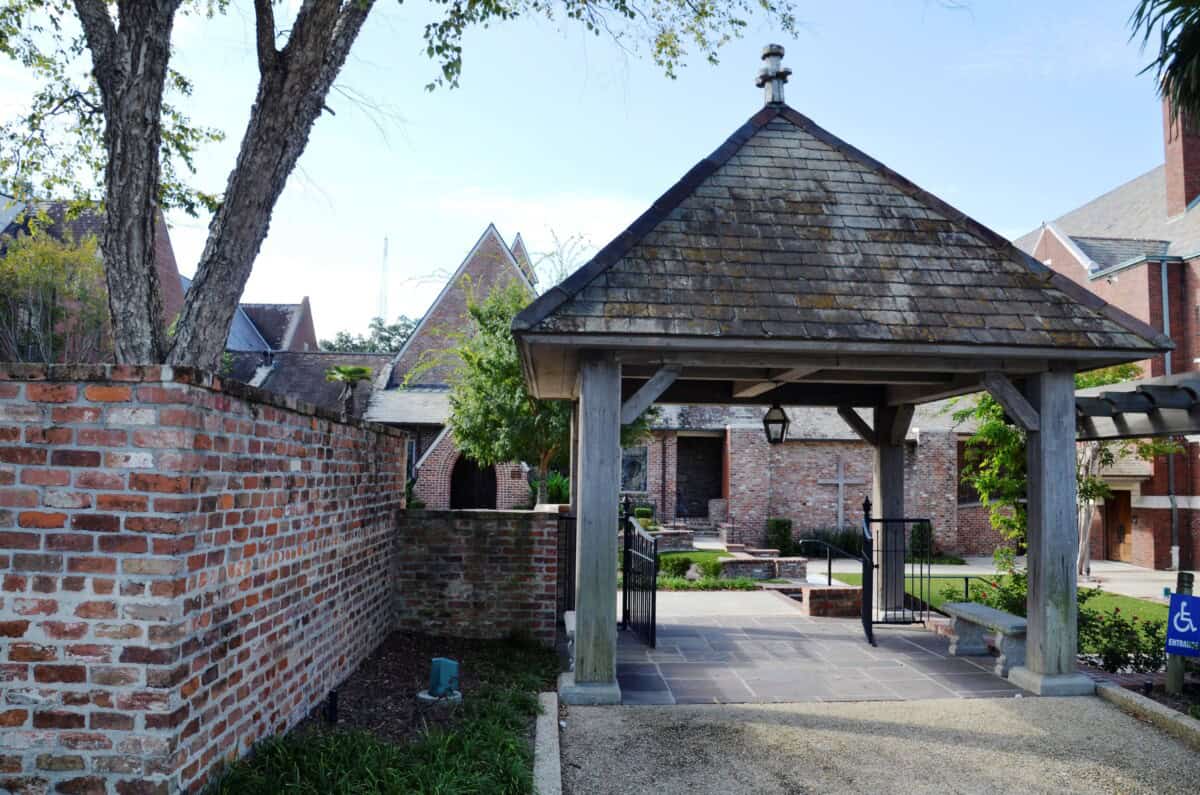 A brick courtyard with a wooden gazebo structure, surrounded by trees and buildings with pitched roofs in the background.