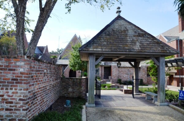 A brick courtyard with a wooden gazebo structure, surrounded by trees and buildings with pitched roofs in the background.