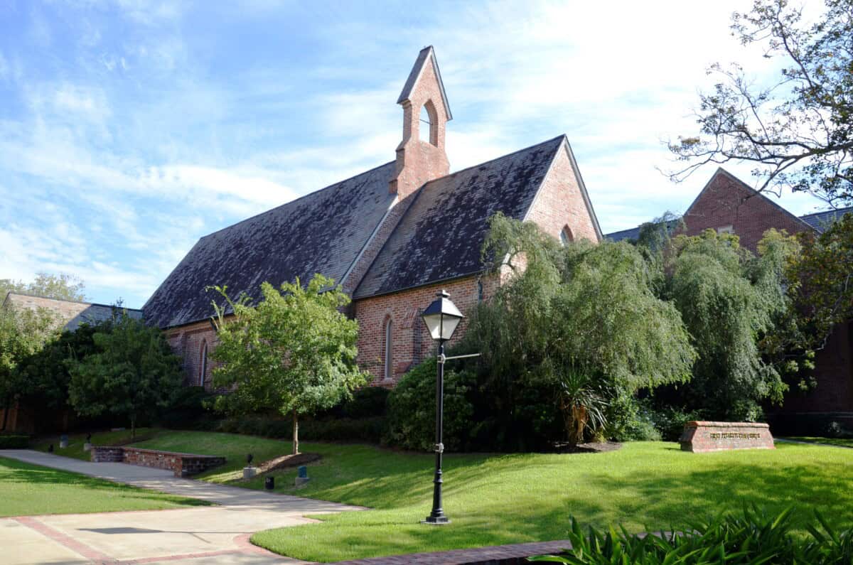 A brick church with a small steeple stands surrounded by green trees, grass, and a streetlamp on a sunny day.