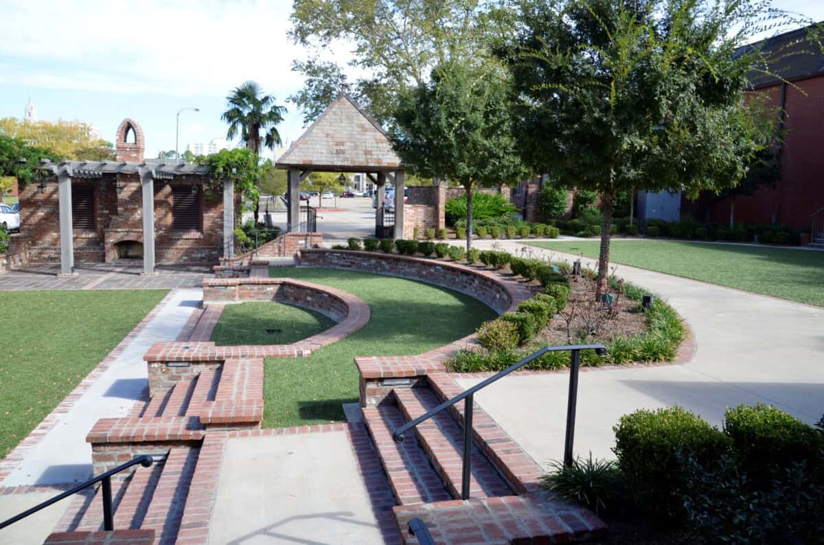 A small outdoor amphitheater with curved brick seating, a grassy area, and surrounding trees and shrubs on a sunny day.