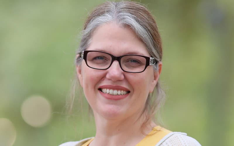 Woman with gray hair and glasses smiling outdoors, dressed in yellow, exudes the warmth of caring Elders against a blurred green background.