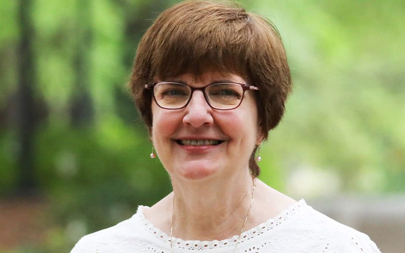 A woman with short brown hair, glasses, and a white top smiles outdoors, perhaps among fellow Elders, with greenery in the background.
