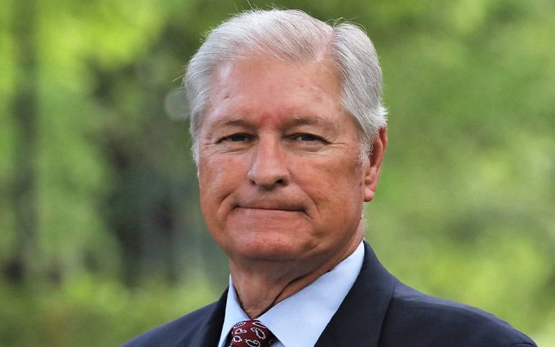Older man with gray hair, dressed in a dark suit and patterned tie, stands outdoors—an image reminiscent of respected elders.