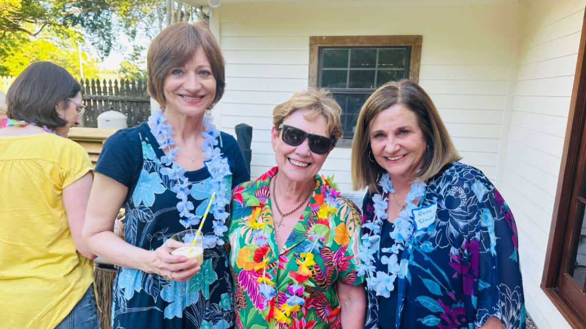 Three women stand outside smiling, wearing floral leis and patterned shirts. One holds a drink, and another wears sunglasses.