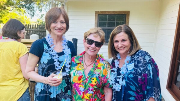 Three women stand outside smiling, wearing floral leis and patterned shirts. One holds a drink, and another wears sunglasses.