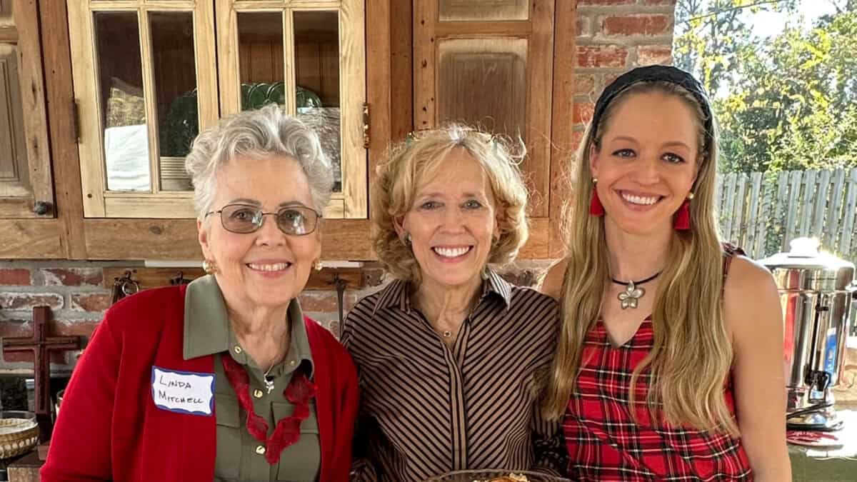 Three women stand indoors, smiling at the camera. One wears a name tag, and a kitchen and wooden cabinets are visible in the background.