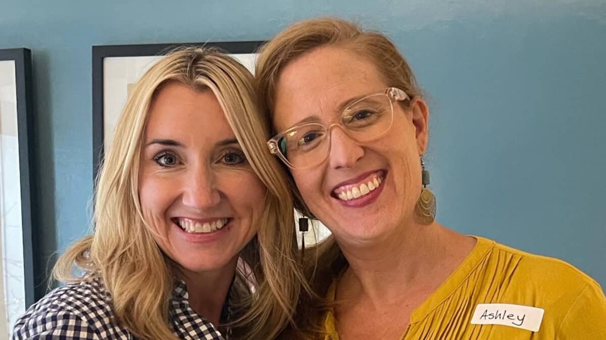 Two women smiling and posing together in front of a blue wall; one wears glasses and a yellow shirt with a nametag reading "Ashley.