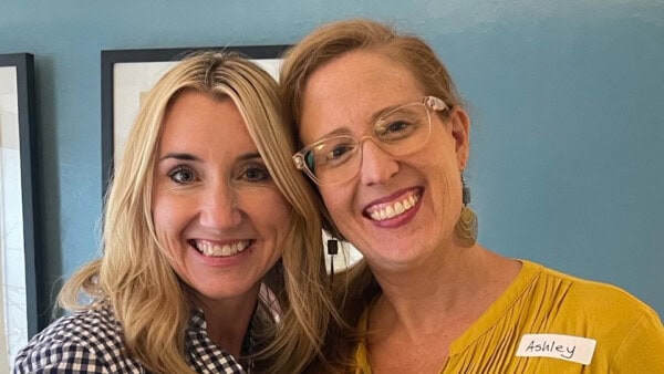 Two women smiling and posing together in front of a blue wall; one wears glasses and a yellow shirt with a nametag reading 