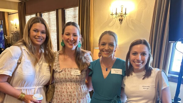 Four women stand together indoors, all smiling at the camera and wearing name tags, with warm lighting in the background.