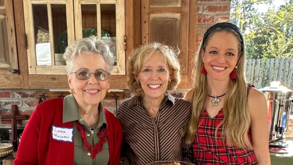 Three women stand indoors, smiling at the camera. One wears a name tag, and a kitchen and wooden cabinets are visible in the background.