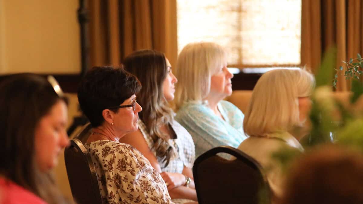 Several women sit in a row at an indoor event, facing forward and listening attentively, with warm lighting in the room.