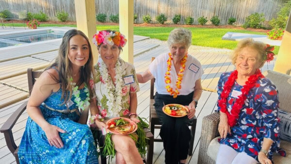 Four women sit outdoors by a pool, wearing leis and floral outfits, holding plates of food, and smiling at the camera.