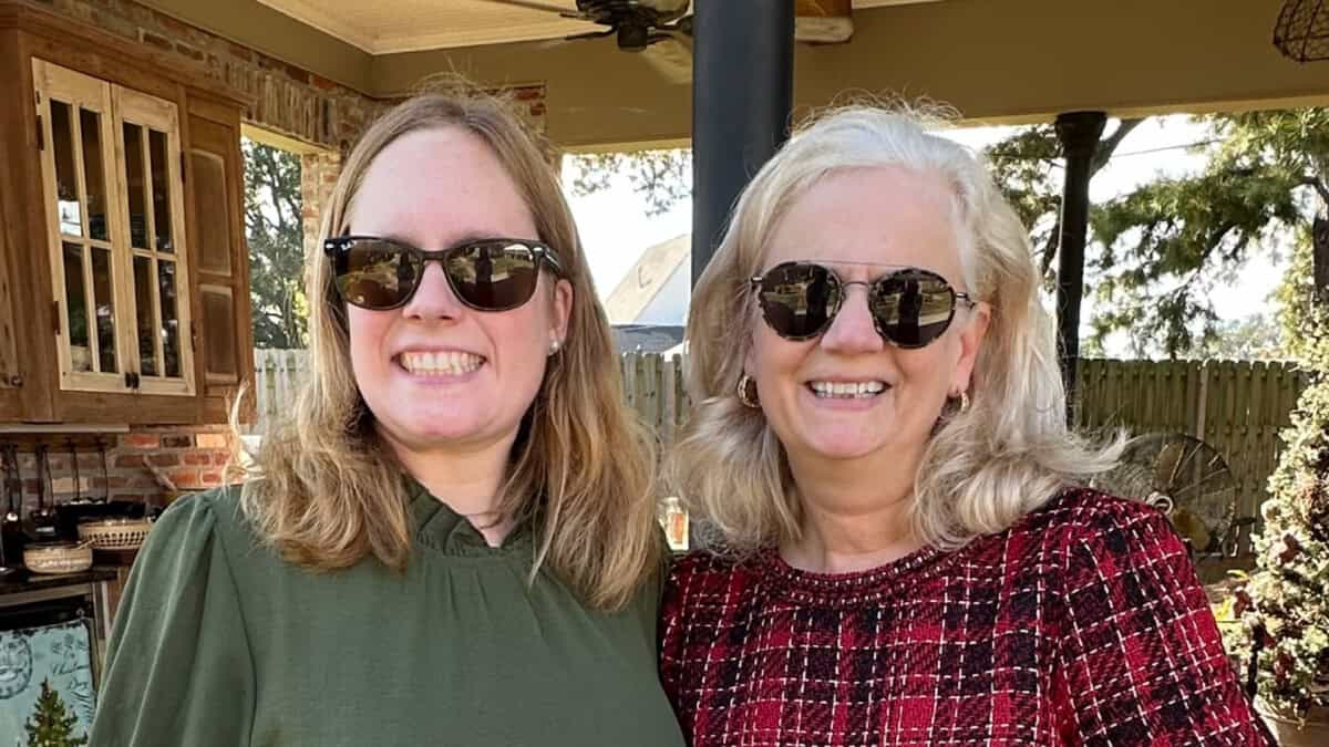 Two women wearing sunglasses stand side by side outdoors, smiling at the camera. There is a wooden fence and greenery in the background.