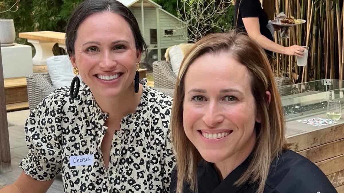 Two women smiling at a table outdoors; one wears a floral top and name tag, the other wears black. Food and drinks are on the table.