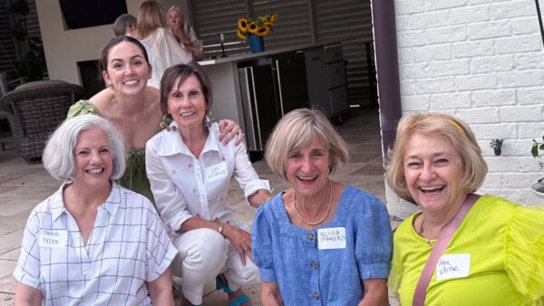Five women, four sitting and one kneeling behind, smile at an outdoor gathering; all wear name tags.