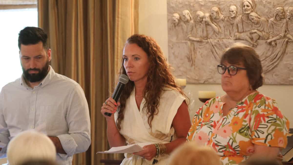 Three adults stand indoors, one holding a microphone and speaking, while the others hold papers; a relief artwork hangs behind them.
