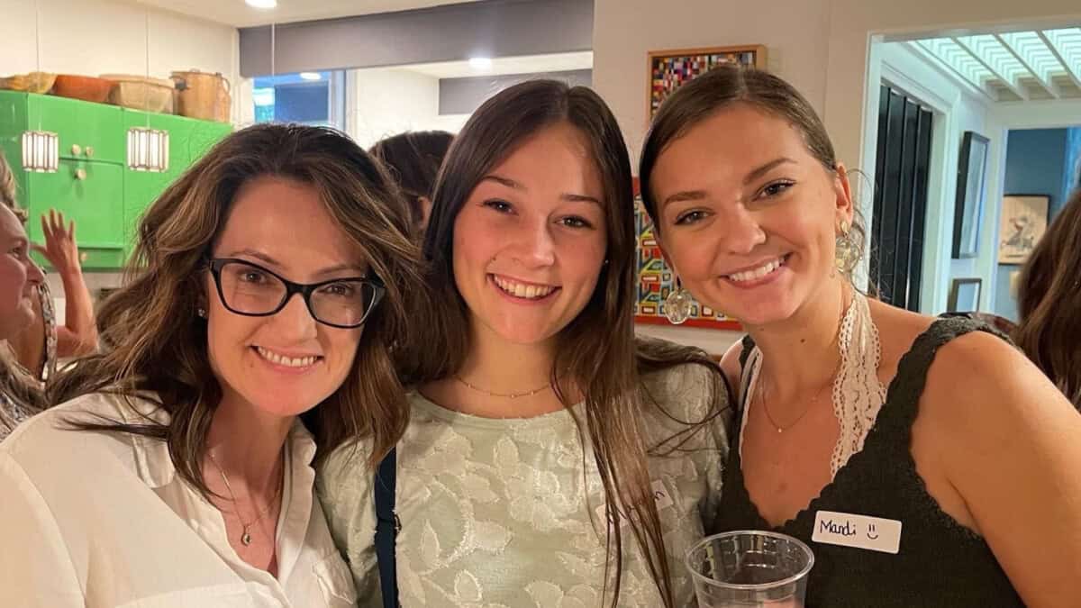 Three women smiling together indoors, one wearing glasses, one holding a drink, and one wearing a name tag that says "Madi.