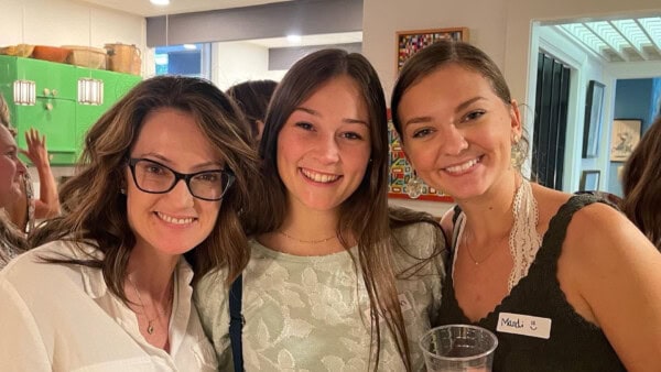 Three women smiling together indoors, one wearing glasses, one holding a drink, and one wearing a name tag that says 