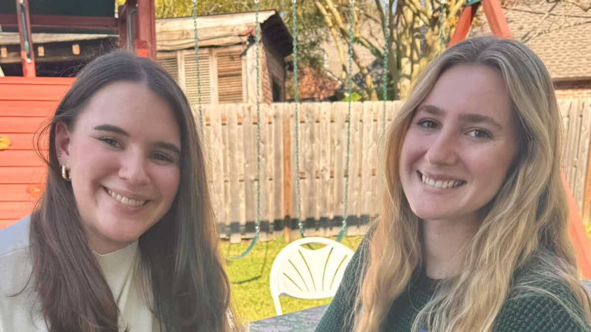 Two young women sit outside at a table, smiling at the camera with a wooden fence and playground equipment in the background.