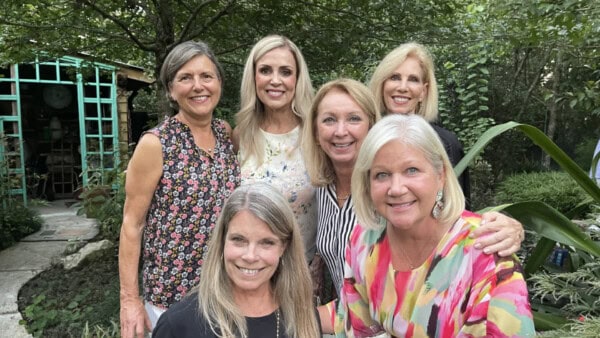 Six women standing and smiling together outdoors in a garden with trees and greenery in the background.