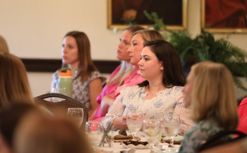 A group of women sit at a table, attentively listening during an event or meeting, with glasses and water bottles in front of them.